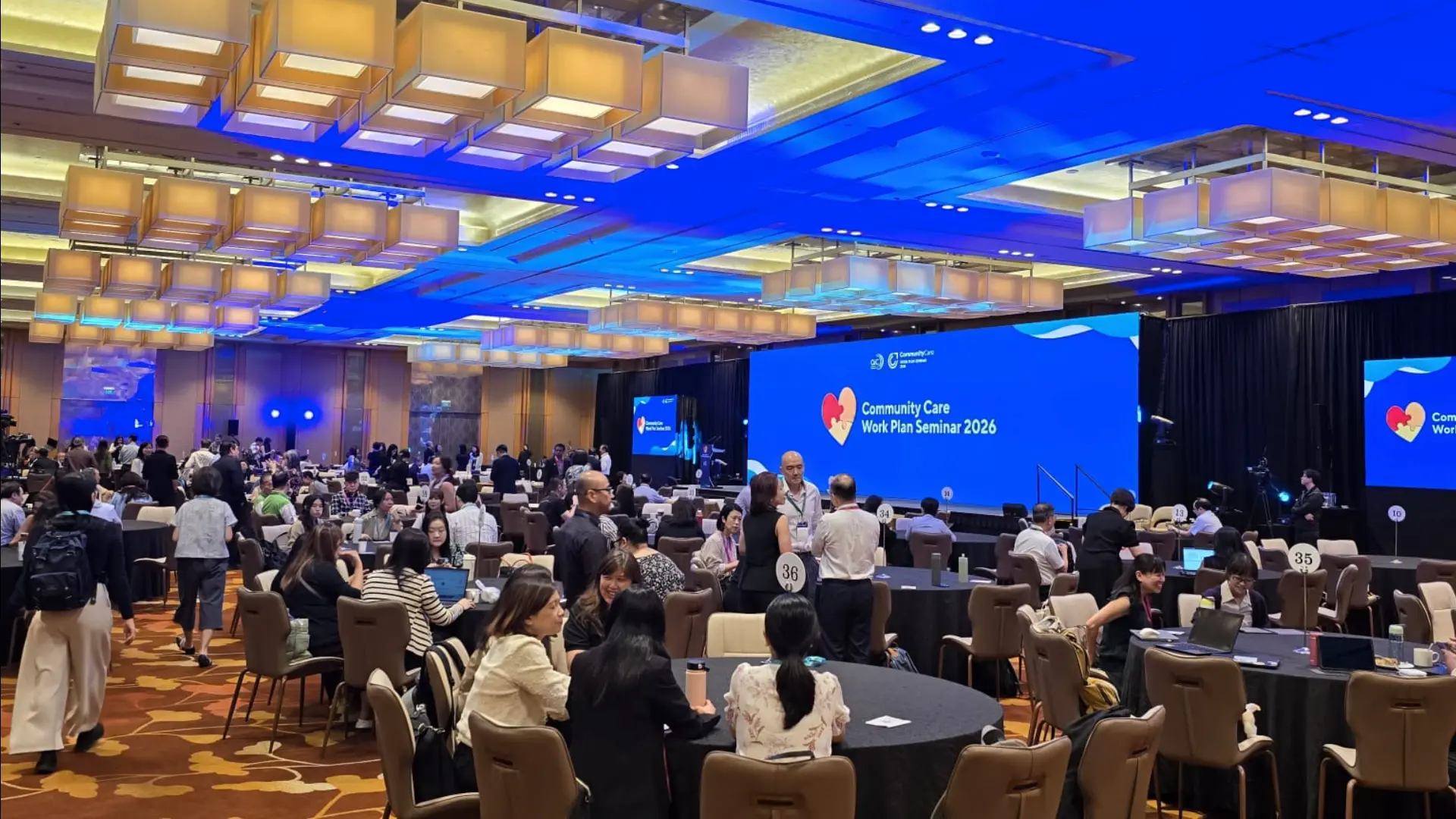 Full ballroom plenary session with blue stage lighting and a large LED backdrop
