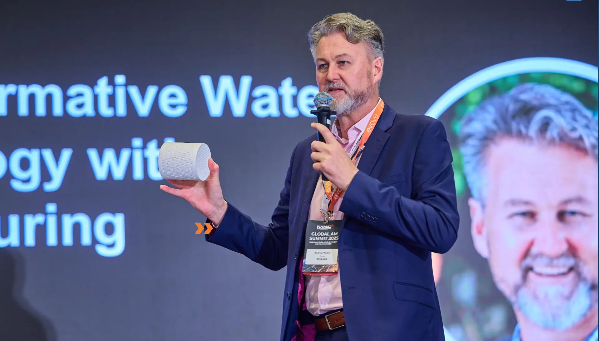Presenter speaking on stage with live camera feed projected beside him