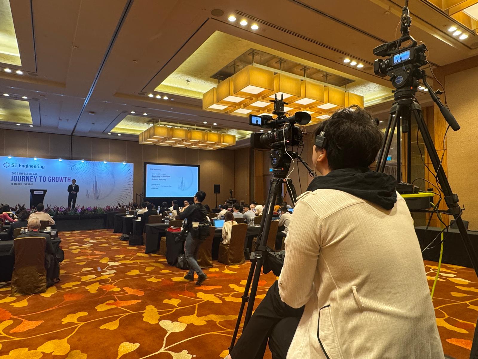 Camera operator filming an investor day presentation from the rear of the ballroom